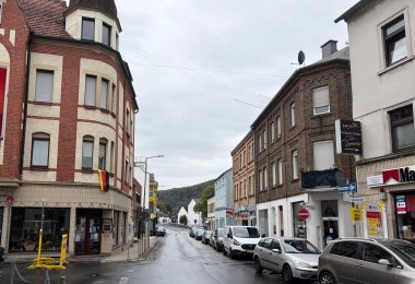 Heutige Ansicht einer Straße in Lahnstein mit mehrgeschossigen Wohn- und Geschäftshäusern auf beiden Seiten. Entlang des rechten Straßenrandes stehen mehrere geparkte Autos. Die Straße führt leicht abwärts und anschließend in einer Kurve weiter. Im Hintergrund ist ein bewaldeter Hang zu sehen.