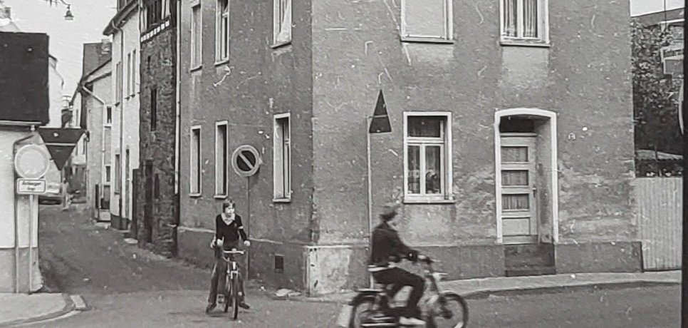 Historische Schwarz-Weiß-Aufnahme einer Straßenecke in Lahnstein. Im Vordergrund fährt ein Mann auf einem Motorrad über die Straße, daneben steht ein Junge mit einem Fahrrad. Hinter einem mehrgeschossigen Wohnhaus erhebt sich ein massiver steinerner Turm. Links führt eine schmale Gasse zwischen den Häusern entlang.