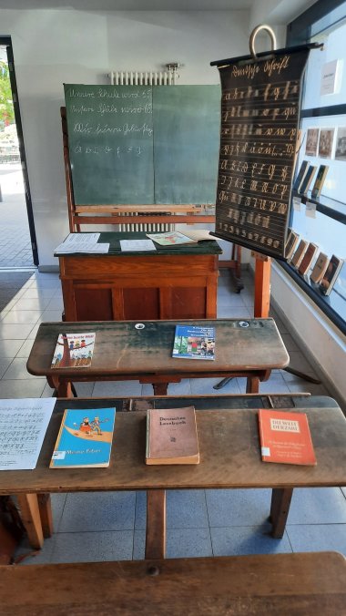 Ein Blick in vergangene Zeiten Blick auf eine historische Schulausstellung mit mehreren alten Holzbänken, einem Lehrerpult und einer großen grünen Schultafel. Auf den Bänken und dem Pult liegen verschiedene Schulbücher und Schriftstücke. Rechts steht eine historische Schautafel mit unterschiedlichen Schreibschriften und Buchstaben. An der grünen Tafel sind mit Kreide Schriftzüge und einzelne Buchstaben zu sehen.
