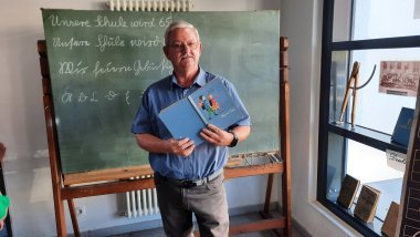 Dieter Halbig, ehemaliger Hausmeister der Schillerschule, schaute sich um und entdeckte vieles aus der eigenen Schulzeit an der Volksschule Niederlahnstein wieder. Ein Mann steht vor einer historischen grünen Schultafel und hält zwei ältere Schulbücher beziehungsweise Fibeln in den Händen. Auf der Tafel befinden sich mit Kreide geschriebene Sätze und Buchstaben. Rechts sind in einem Fenster weitere historische Bücher und ein altes Klassenfoto ausgestellt.