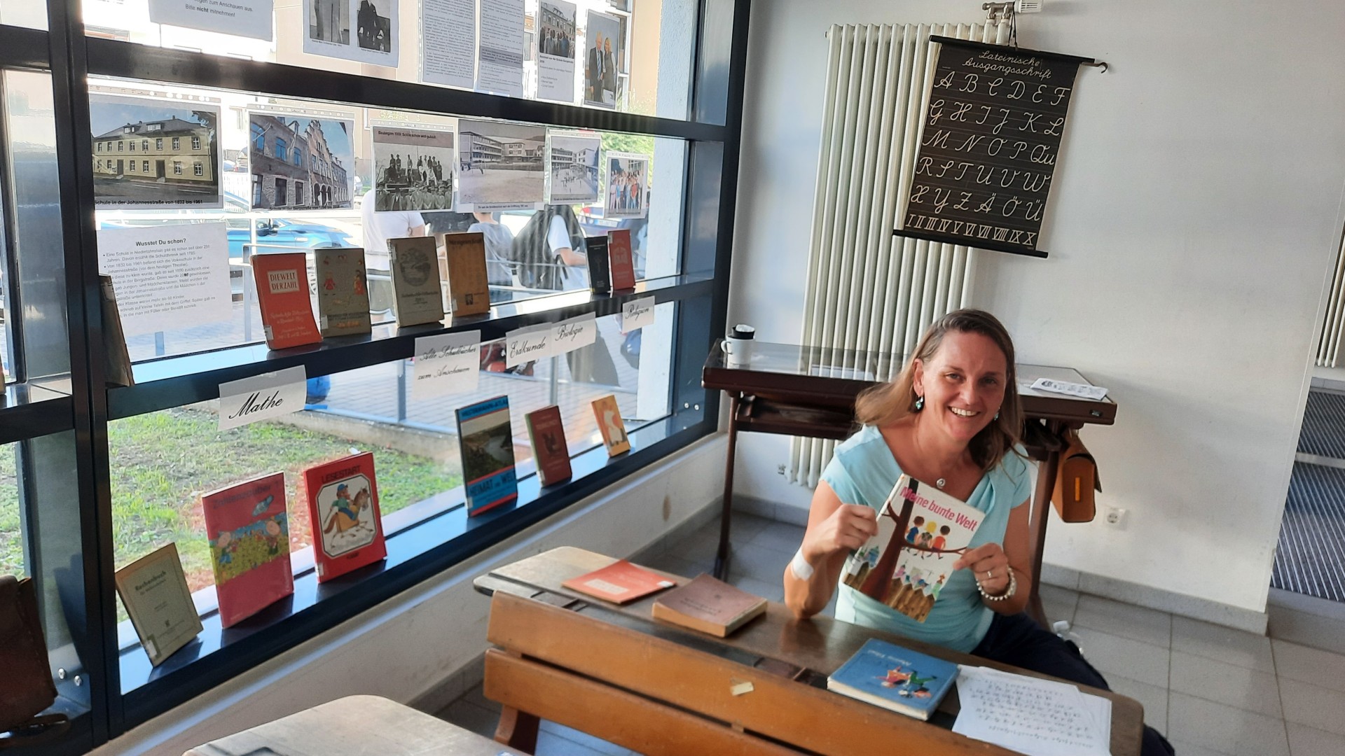 Eine Frau sitzt an einer historischen Schulbank und hält das Schulbuch „Meine bunte Welt“ in den Händen. Auf der Schulbank liegen weitere ältere Bücher und ein beschriebenes Blatt. Im Fenster hinter ihr sind zahlreiche historische Schulbücher, Fotografien und Infor