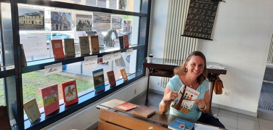Auch Frau Zickhardt, Lehrkraft an der Schillerschule, entdeckte ihre eigene Schulfibel. Eine Frau sitzt an einer historischen Schulbank und hält das Schulbuch „Meine bunte Welt“ in den Händen. Auf der Schulbank liegen weitere ältere Bücher und ein beschriebenes Blatt. Im Fenster hinter ihr sind zahlreiche historische Schulbücher, Fotografien und Infor