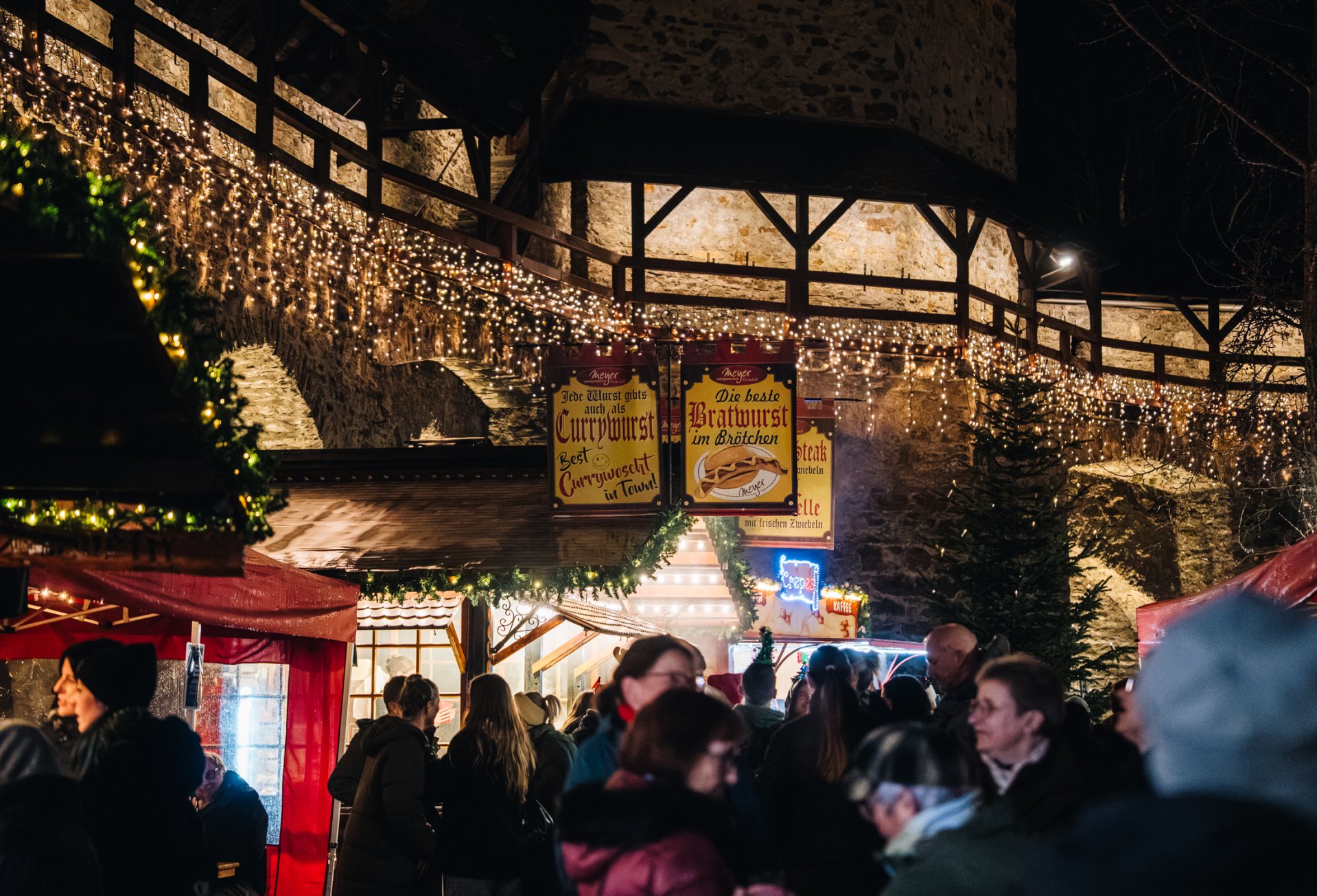 Abendliche Aufnahme eines gut besuchten Weihnachtsmarktes vor einer historischen Steinmauer. Zahlreiche Lichterketten schmücken die Mauer und die Verkaufsstände. Im Vordergrund stehen Besucherinnen und Besucher zwischen rot überdachten und weihnachtlich dekorierten Marktständen. Im Hintergrund sind beleuchtete Werbeschilder an einem Imbissstand sowie mehrere Tannenbäume zu sehen.