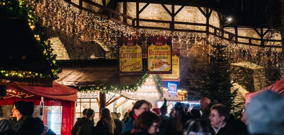 Nikolausmarkt Lahnstein Abendliche Aufnahme eines gut besuchten Weihnachtsmarktes vor einer historischen Steinmauer. Zahlreiche Lichterketten schmücken die Mauer und die Verkaufsstände. Im Vordergrund stehen Besucherinnen und Besucher zwischen rot überdachten und weihnachtlich dekorierten Marktständen. Im Hintergrund sind beleuchtete Werbeschilder an einem Imbissstand sowie mehrere Tannenbäume zu sehen.