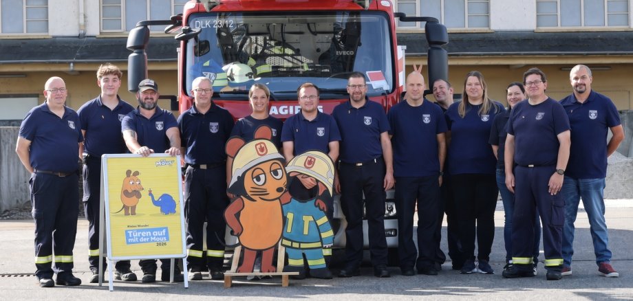 Türen auf mit der Maus Gruppenfoto von Mitgliedern der Feuerwehr Lahnstein vor einem roten Feuerwehrfahrzeug mit Drehleiter. Vor der Gruppe stehen ein Aufsteller zur Aktion „Türen auf mit der Maus 2026“ sowie zwei große Figuren der Maus in Feuerwehrkleidung.