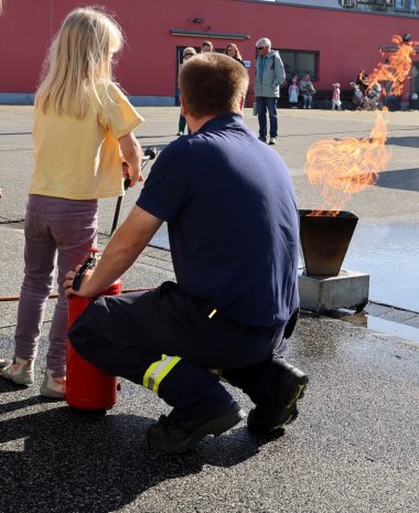 Türen auf mit der Maus Ein Feuerwehrmann kniet neben einem Feuerlöscher und begleitet ein Kind bei einer Löschübung. Vor den beiden brennt ein Feuer in einem Metallbehälter. Im Hintergrund stehen mehrere Personen vor einem Gebäude und beobachten die Übung.