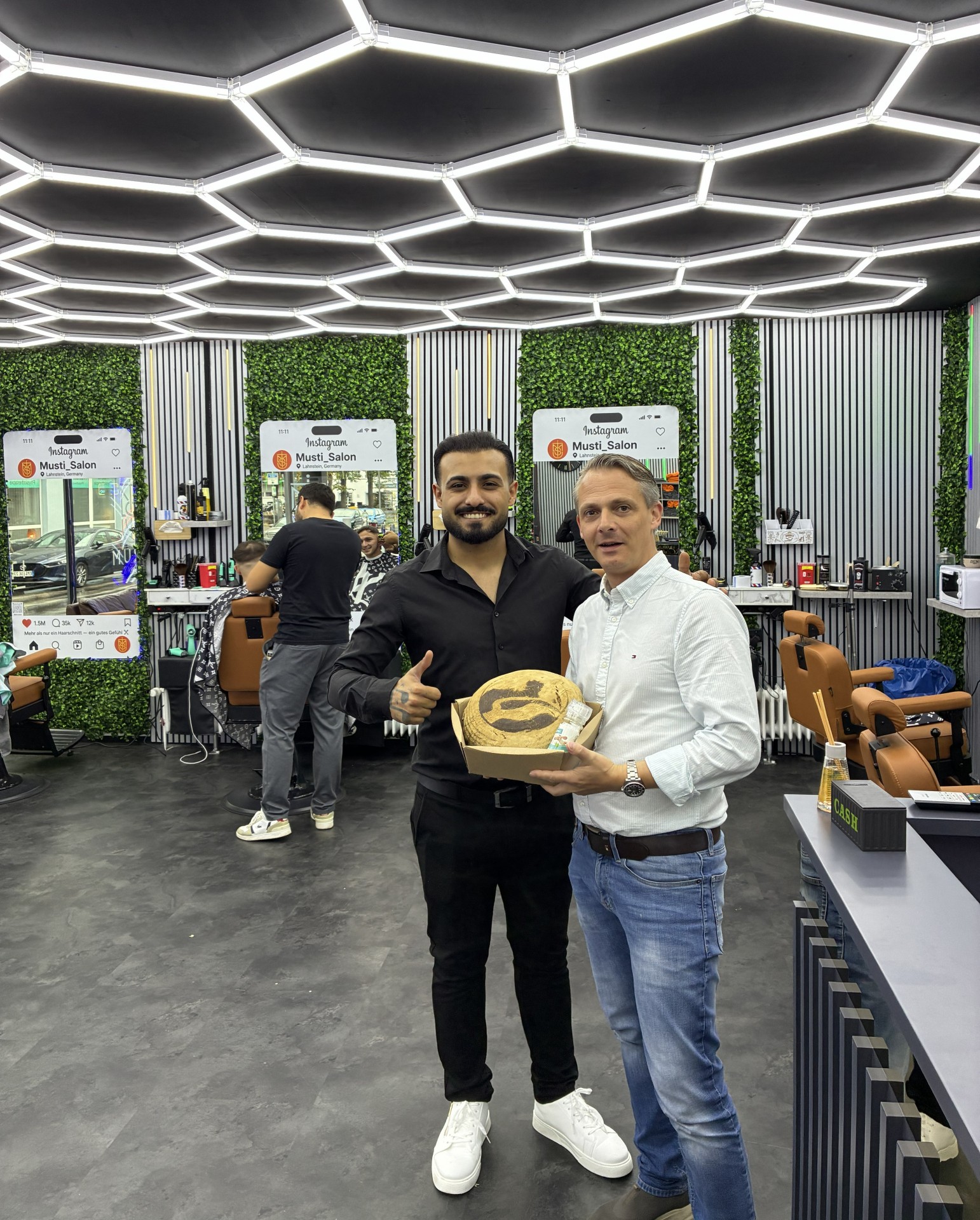 Zwei Männer stehen in einem modern eingerichteten Friseursalon nebeneinander. Einer hält einen Karton mit einem Brot und weiteren kleinen Präsenten in den Händen. Im Hintergrund sind Friseurstühle, Spiegel, Arbeitsplätze sowie begrünte Wandflächen zu sehen.