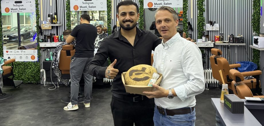 Zwei Männer stehen in einem modern eingerichteten Friseursalon nebeneinander. Einer hält einen Karton mit einem Brot und weiteren kleinen Präsenten in den Händen. Im Hintergrund sind Friseurstühle, Spiegel, Arbeitsplätze sowie begrünte Wandflächen zu sehen.