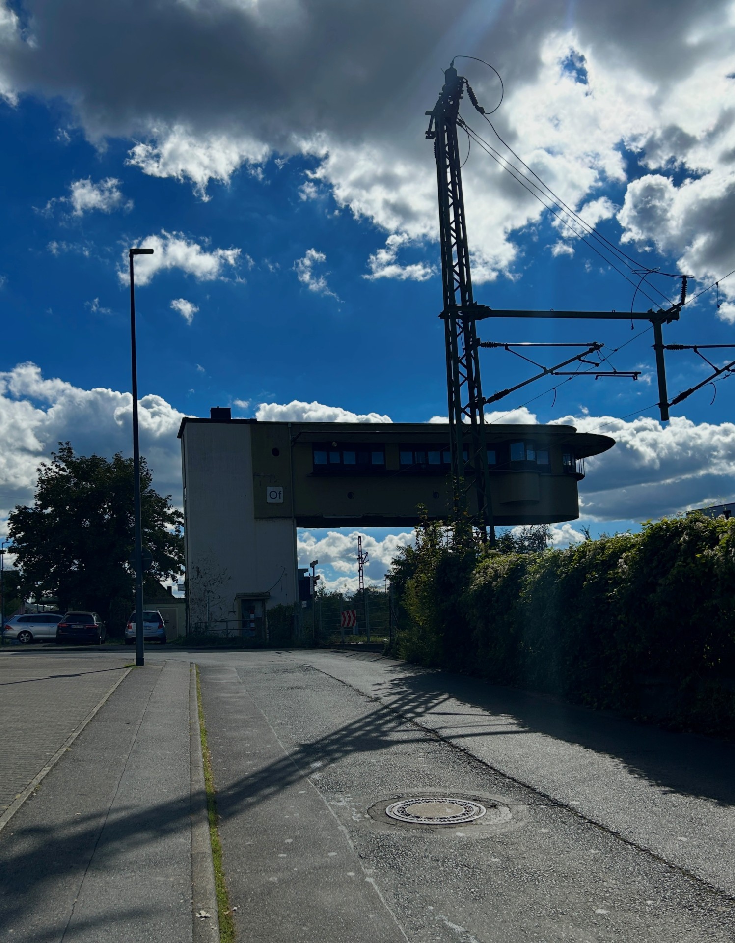 Blick auf das Stellwerk an der Bürgermeister-Müller-Straße in Lahnstein. Im Vordergrund verläuft die Straße mit angrenzendem Gehweg. Rechts ragen ein Oberleitungsmast und Stromleitungen der Bahn in den Himmel. Im Hintergrund ist das Stellwerksgebäude mit seinem auskragenden oberen Geschoss zu sehen.