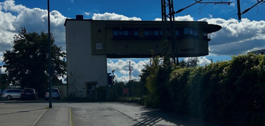 Bürgermeister-Müller-Straße Lahnstein Blick auf das Stellwerk an der Bürgermeister-Müller-Straße in Lahnstein. Im Vordergrund verläuft die Straße mit angrenzendem Gehweg. Rechts ragen ein Oberleitungsmast und Stromleitungen der Bahn in den Himmel. Im Hintergrund ist das Stellwerksgebäude mit seinem auskragenden oberen Geschoss zu sehen.