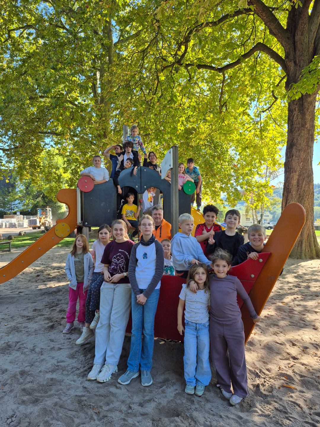 Eine Gruppe von Kindern und mehreren Erwachsenen posiert auf und vor einem großen Spielgerät mit Rutschen auf einem sandigen Spielplatz. Im Hintergrund spenden große Bäume mit grünem und gelblichem Herbstlaub Schatten.
