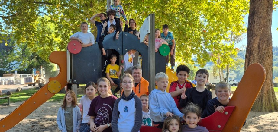 Herbstfreizeit im Jugendkulturzentrum Lahnstein Eine Gruppe von Kindern und mehreren Erwachsenen posiert auf und vor einem großen Spielgerät mit Rutschen auf einem sandigen Spielplatz. Im Hintergrund spenden große Bäume mit grünem und gelblichem Herbstlaub Schatten.