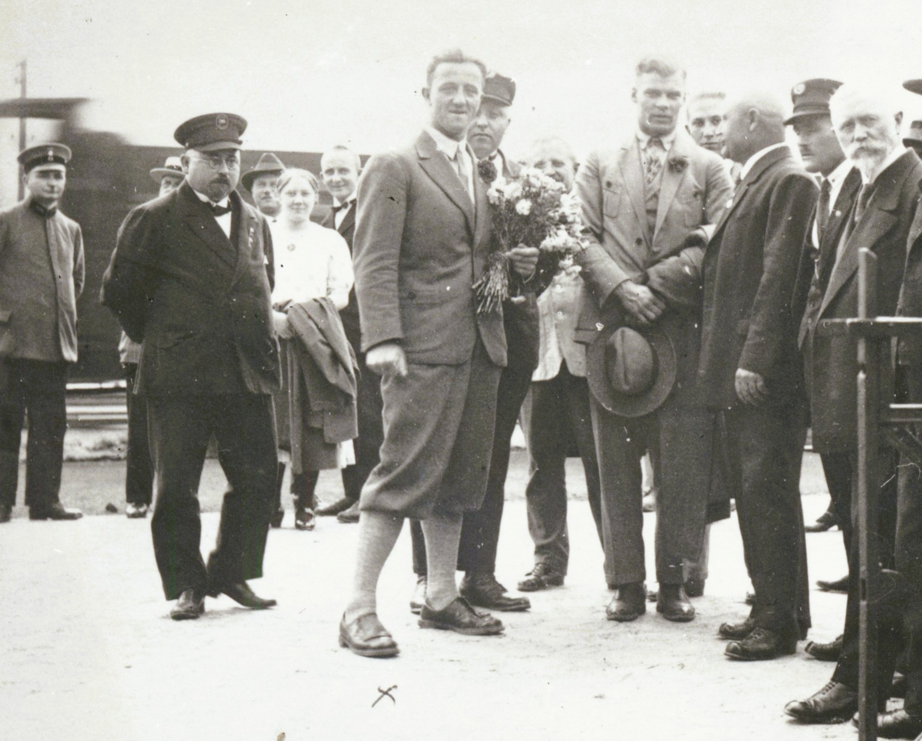 Historische Schwarz-Weiß-Aufnahme einer Gruppe festlich gekleideter Männer und Frauen an einem Bahnhof. Im Mittelpunkt steht ein junger Mann im Anzug, neben ihm eine Person mit einem Blumenstrauß. Im Hintergrund sind weitere Personen und ein Eisenbahnwaggon zu sehen.