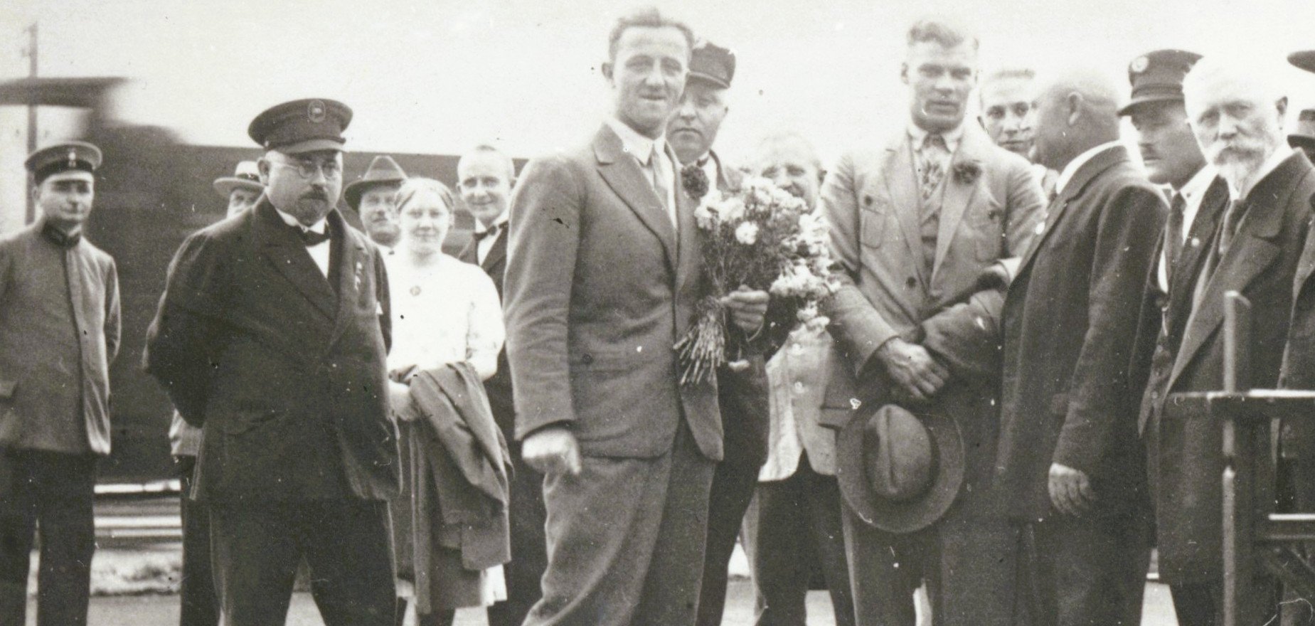 Empfang von Schwimmweltmeister Erich Rademacher (in Knickerbockerhose) in Oberlahnstein mit Blumen Historische Schwarz-Weiß-Aufnahme einer Gruppe festlich gekleideter Männer und Frauen an einem Bahnhof. Im Mittelpunkt steht ein junger Mann im Anzug, neben ihm eine Person mit einem Blumenstrauß. Im Hintergrund sind weitere Personen und ein Eisenbahnwaggon zu sehen.