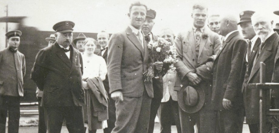 Empfang von Schwimmweltmeister Erich Rademacher (in Knickerbockerhose) in Oberlahnstein mit Blumen Historische Schwarz-Weiß-Aufnahme einer Gruppe festlich gekleideter Männer und Frauen an einem Bahnhof. Im Mittelpunkt steht ein junger Mann im Anzug, neben ihm eine Person mit einem Blumenstrauß. Im Hintergrund sind weitere Personen und ein Eisenbahnwaggon zu sehen.