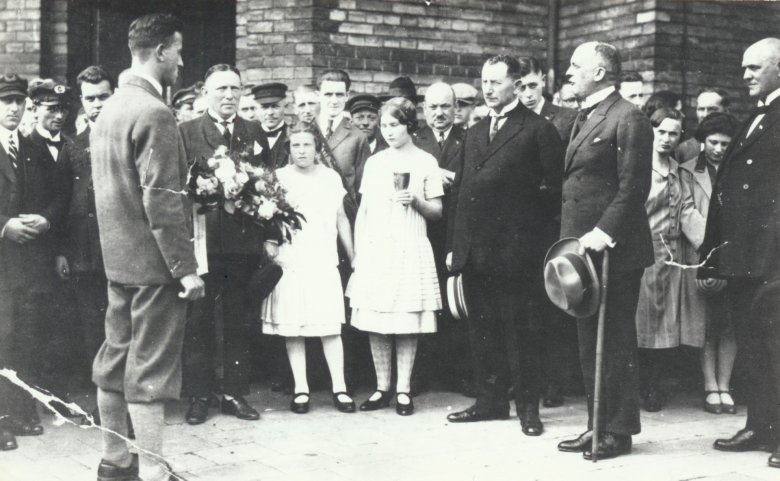 Empfang von Schwimmweltmeister Erich Rademacher (links vorne mit Knickerbockerhose) in Niederlahnstein mit Weinpokal Historische Schwarz-Weiß-Aufnahme eines feierlichen Empfangs vor einem Backsteingebäude. Zahlreiche Männer in Anzügen sowie Frauen und Kinder haben sich versammelt. Im Vordergrund steht ein junger Mann im Anzug, dem sich die Gruppe zuwendet. Zwei Mädchen in hellen Kleidern stehen mit einem Blumenstrauß und einem Pokal in der Mitte.