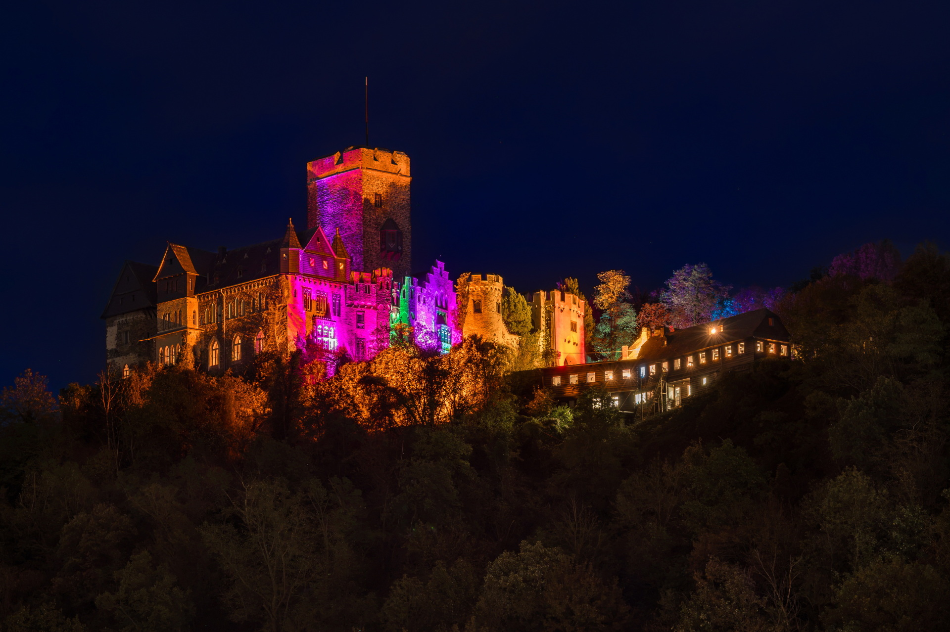 Nächtliche Gesamtansicht der Burg Lahneck in Lahnstein, die auf einer bewaldeten Anhöhe liegt. Die mittelalterliche Burganlage mit ihren hohen Mauern, Zinnen, Türmen und steilen Dächern wird beim Lichtfestival rheinleuchten in verschiedenen Farben beleuchtet. Der große viereckige Hauptturm leuchtet in Pink und Orange, während die angrenzenden Gebäudefassaden in kräftigem Pink, Grün und warmem Gelborange erstrahlen. Rechts schließen sich weitere beleuchtete Türme und ein lang gestrecktes Gebäude mit hellen Fenstern an. Unterhalb der Burg sind die dunklen Umrisse zahlreicher Bäume zu sehen. Darüber erstreckt sich ein tief dunkelblauer Nachthimmel.