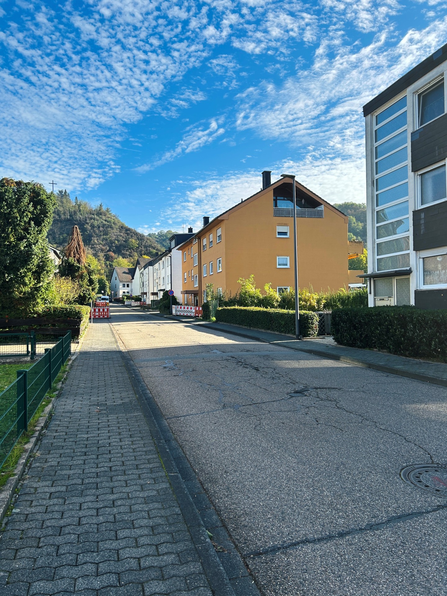 Blick auf eine Wohnstraße in Lahnstein bei sonnigem Wetter. Die asphaltierte Fahrbahn weist zahlreiche Risse auf und führt geradeaus zwischen mehrstöckigen Wohnhäusern hindurch. Auf der rechten Seite stehen ein großes ockerfarbenes Wohnhaus mit Satteldach und Balkonen sowie ein modernes Gebäude mit grauer Fassade und großen Fensterflächen. Links verläuft ein gepflasterter Gehweg entlang eines niedrigen grünen Metallzauns und dichter Bäume und Sträucher. Weiter hinten sind auf beiden Straßenseiten rot-weiße Baustellenabsperrungen aufgestellt. Im Hintergrund erheben sich bewaldete Hügel. Der Himmel ist leuchtend blau und von zahlreichen kleinen weißen Wolken bedeckt.