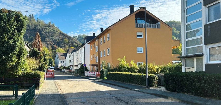 Josef-Rätz-Straße Blick auf eine Wohnstraße in Lahnstein bei sonnigem Wetter. Die asphaltierte Fahrbahn weist zahlreiche Risse auf und führt geradeaus zwischen mehrstöckigen Wohnhäusern hindurch. Auf der rechten Seite stehen ein großes ockerfarbenes Wohnhaus mit Satteldach und Balkonen sowie ein modernes Gebäude mit grauer Fassade und großen Fensterflächen. Links verläuft ein gepflasterter Gehweg entlang eines niedrigen grünen Metallzauns und dichter Bäume und Sträucher. Weiter hinten sind auf beiden Straßenseiten rot-weiße Baustellenabsperrungen aufgestellt. Im Hintergrund erheben sich bewaldete Hügel. Der Himmel ist leuchtend blau und von zahlreichen kleinen weißen Wolken bedeckt.