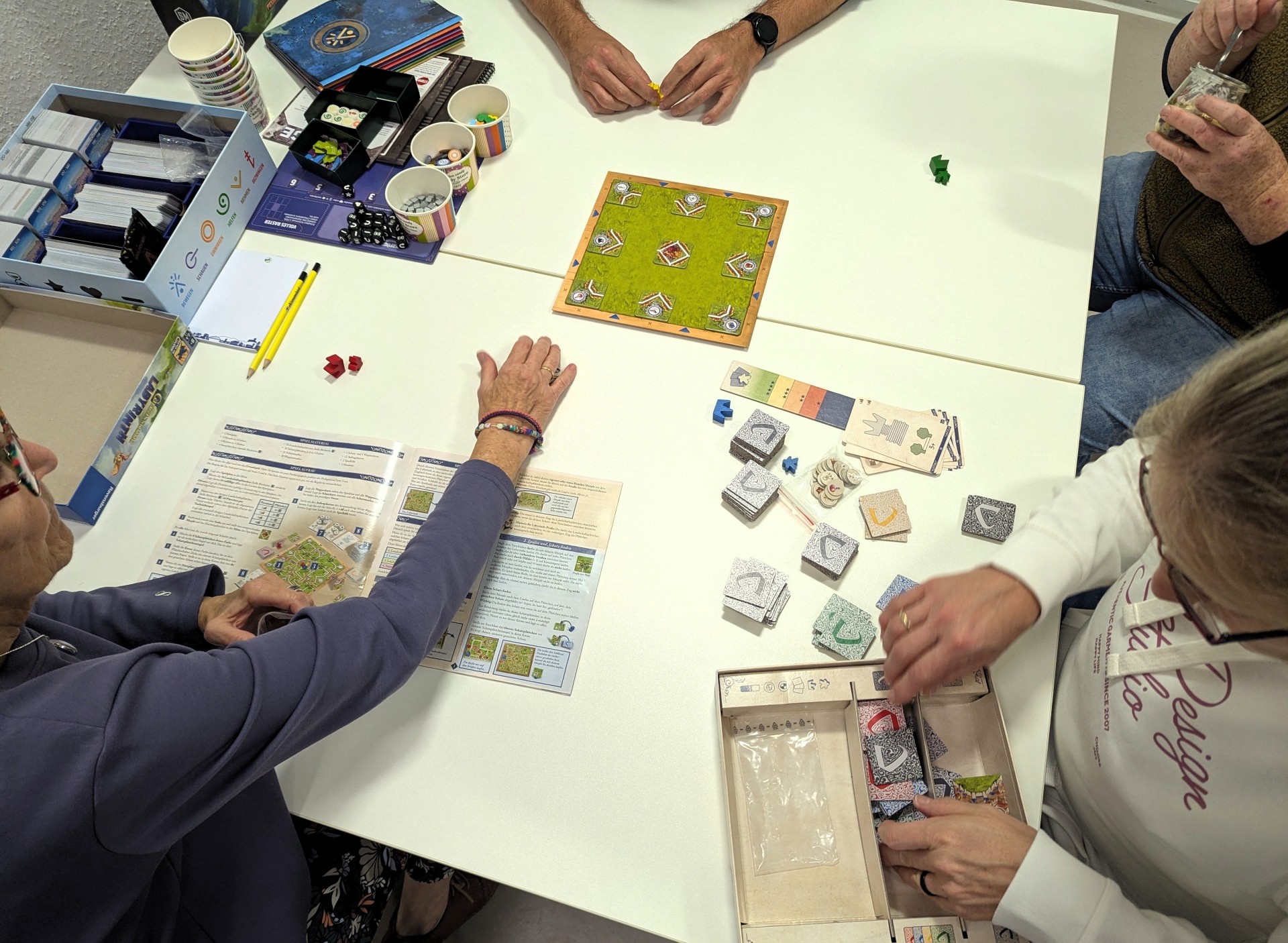 Vier Personen sitzen beim Spieleabend in der Stadtbücherei Lahnstein an einem Tisch und spielen gemeinsam ein Brettspiel. Auf dem Tisch liegen ein Spielbrett, Karten, Würfel, Spielfiguren und Spielanleitungen.