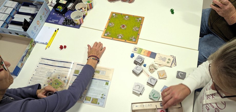 Spieleabend Stadtbücherei Vier Personen sitzen beim Spieleabend in der Stadtbücherei Lahnstein an einem Tisch und spielen gemeinsam ein Brettspiel. Auf dem Tisch liegen ein Spielbrett, Karten, Würfel, Spielfiguren und Spielanleitungen.
