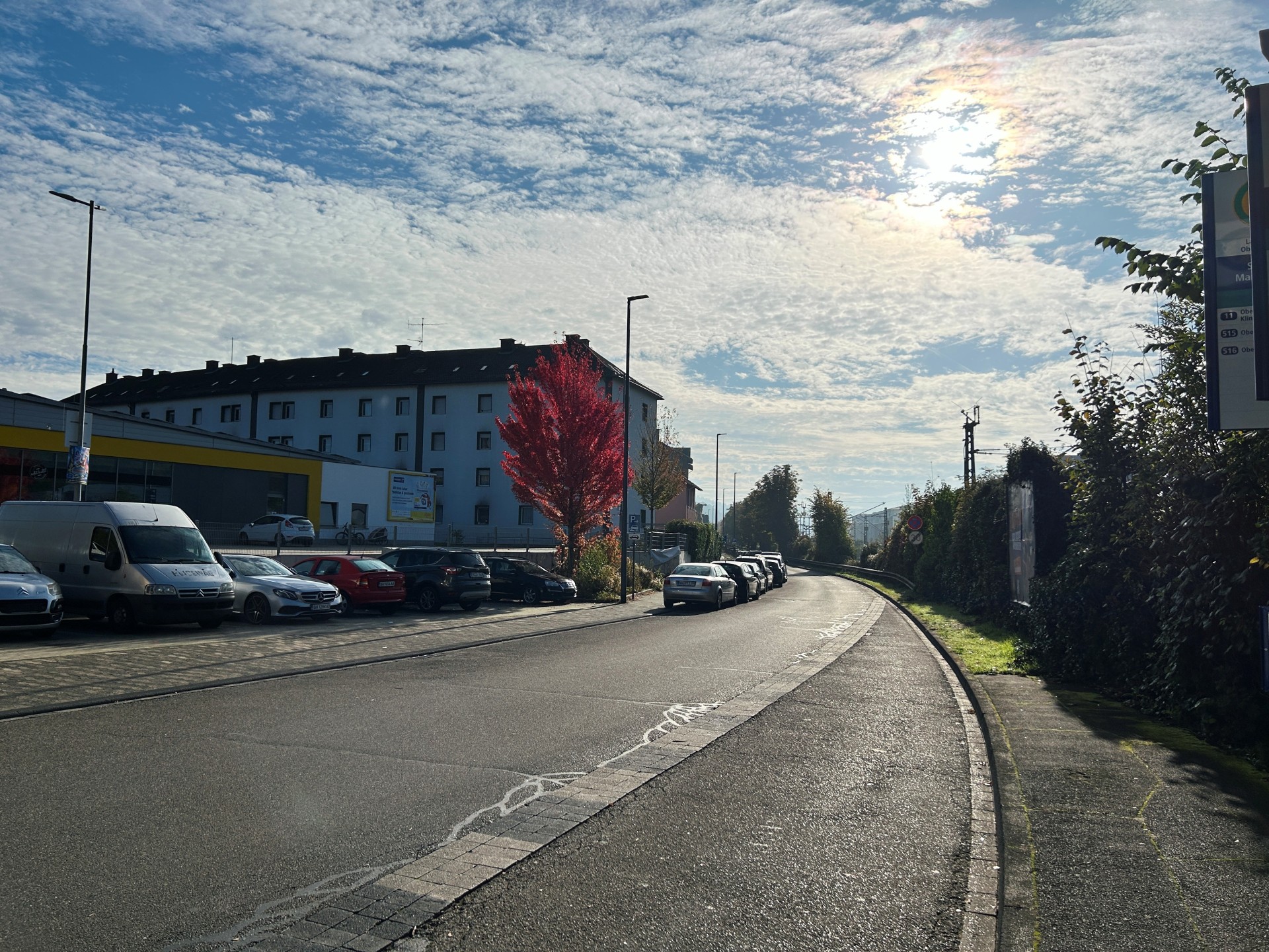 Tagesaufnahme der Braubacher Straße in Lahnstein. Die asphaltierte Straße verläuft in einer leichten Rechtskurve und wird auf beiden Seiten von Gehwegen begleitet. Links stehen zahlreiche geparkte Autos vor einem mehrstöckigen Wohngebäude mit heller Fassade und einem flachen Gewerbegebäude mit gelben Fassadenelementen. Ein Baum mit leuchtend roten Herbstblättern fällt zwischen den Gebäuden und Fahrzeugen besonders auf. Rechts verläuft ein schmaler Gehweg entlang einer hohen, dicht bewachsenen Hecke. Im Hintergrund sind Oberleitungsmasten der Bahn zu erkennen. Über der Straße erstreckt sich ein blauer Himmel mit zahlreichen kleinen Wolken, durch die die Sonne scheint.