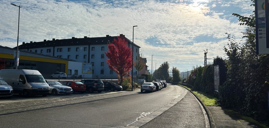 Tagesaufnahme der Braubacher Straße in Lahnstein. Die asphaltierte Straße verläuft in einer leichten Rechtskurve und wird auf beiden Seiten von Gehwegen begleitet. Links stehen zahlreiche geparkte Autos vor einem mehrstöckigen Wohngebäude mit heller Fassade und einem flachen Gewerbegebäude mit gelben Fassadenelementen. Ein Baum mit leuchtend roten Herbstblättern fällt zwischen den Gebäuden und Fahrzeugen besonders auf. Rechts verläuft ein schmaler Gehweg entlang einer hohen, dicht bewachsenen Hecke. Im Hintergrund sind Oberleitungsmasten der Bahn zu erkennen. Über der Straße erstreckt sich ein blauer Himmel mit zahlreichen kleinen Wolken, durch die die Sonne scheint.