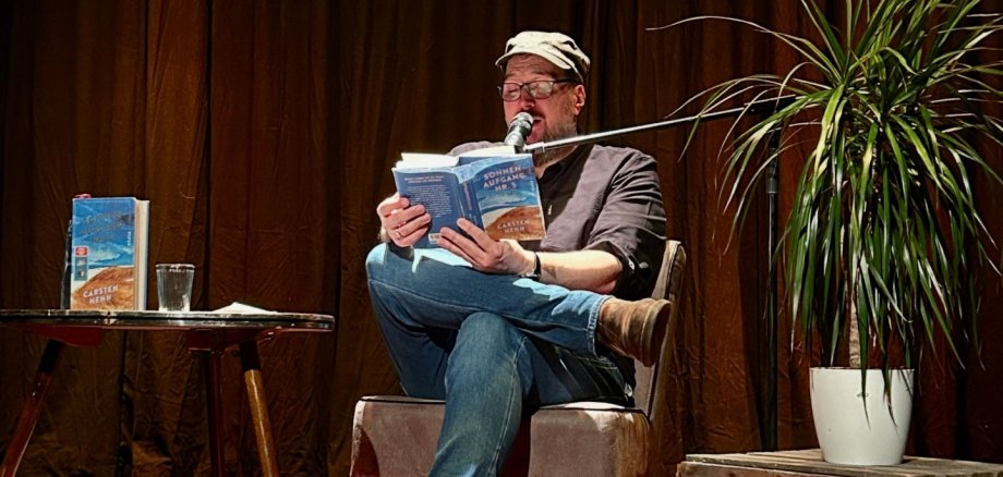 Lesung Carsten Henn Der Autor Carsten Henn sitzt mit überschlagenen Beinen auf einem gepolsterten Stuhl auf einer kleinen Bühne und liest aus seinem Buch „Sonnenaufgang Nr. 5“. Er trägt eine helle Schirmmütze, eine Brille, ein dunkles Hemd, eine blaue Jeans und braune Schuhe. Vor seinem Gesicht befindet sich ein Mikrofon an einem Ständer. Links neben ihm steht ein kleiner runder Tisch mit einem aufgestellten Exemplar seines Buches, einem Wasserglas und einigen Papieren. Rechts steht eine große grüne Zimmerpflanze in einem weißen Topf auf einer Holzkiste. Ein großer rot gemusterter Teppich bedeckt den Bühnenboden. Den Hintergrund bildet ein dunkelbrauner Vorhang, während der Autor von warmem Scheinwerferlicht beleuchtet wird.