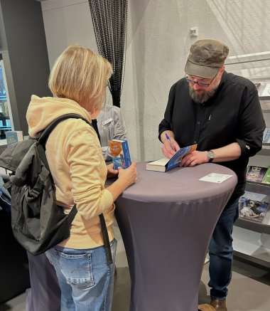 Lesung Carsten Henn Der Autor Carsten Henn steht an einem runden, mit einer grauen Tischhusse bedeckten Stehtisch und signiert mit einem blauen Stift ein aufgeschlagenes Buch. Er trägt eine graubraune Kappe, eine Brille, ein schwarzes Hemd und eine blaue Jeans. Ihm gegenüber steht eine Frau mit schulterlangen blonden Haaren, einem hellgelben Kapuzenpullover, blauer Jeans und schwarzem Rucksack. Sie hält ein weiteres Buch des Autors in den Händen und schaut ihm beim Signieren zu. Im Hintergrund sind eine weitere Person sowie Bücherregale zu sehen.