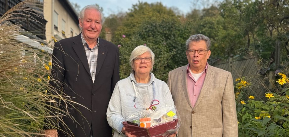 Beigeordneter Manfred Radermacher gratulierte zu 50 Ehejahren. Drei ältere Personen stehen nebeneinander in einem gepflegten Garten und blicken in die Kamera. Links steht ein großer Mann mit kurzen grauen Haaren, der ein dunkles Sakko über einem karierten Hemd und eine blaue Jeans trägt. In der Mitte steht eine Frau mit kurzen weißen Haaren und Brille. Sie trägt einen hellgrauen Kapuzenpullover und eine blaue Jeans und hält einen in durchsichtige Folie verpackten Präsentkorb mit verschiedenen Geschenken in den Händen. Rechts steht ein Mann mit kurzen grauen Haaren und Brille, der ein beigefarbenes Sakko über einem rosafarbenen Hemd und eine blaue Jeans trägt. Die drei Personen stehen auf einer Rasenfläche, umgeben von hohen Ziergräsern, grünen Sträuchern und gelb blühenden Pflanzen. Rechts im Vordergrund sind kleine Tierfiguren als Gartendekoration zu sehen. Im Hintergrund befinden sich ein Wohnhaus, ein Holzzaun und mehrere Bäume.