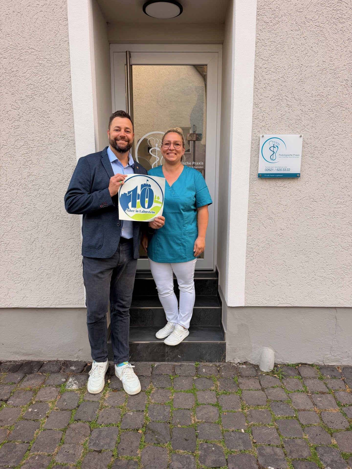 Ein Mann und eine Frau stehen lächelnd vor dem Eingang eines Gebäudes und halten gemeinsam ein rundes Schild mit der Aufschrift „10 Jahre in Lahnstein“ in Blau, Grün und Weiß. Der Mann links trägt ein dunkelblaues Sakko, ein hellblaues Hemd, eine dunkle Hose und weiße Turnschuhe. Er hat kurze dunkle Haare und einen Bart. Die Frau rechts trägt eine türkisfarbene Tunika, eine weiße Hose und weiße Schuhe. Sie hat blonde, zusammengebundene Haare und trägt eine Brille. Hinter den beiden befindet sich eine gläserne Eingangstür mit weißem Rahmen. Rechts daneben hängt an der hell verputzten Hauswand ein Schild mit der Aufschrift „Podologische Praxis“ und einem stilisierten Fuß als Logo. Die beiden stehen auf und vor dunklen Steinstufen, die zu einem gepflasterten Gehweg führen.