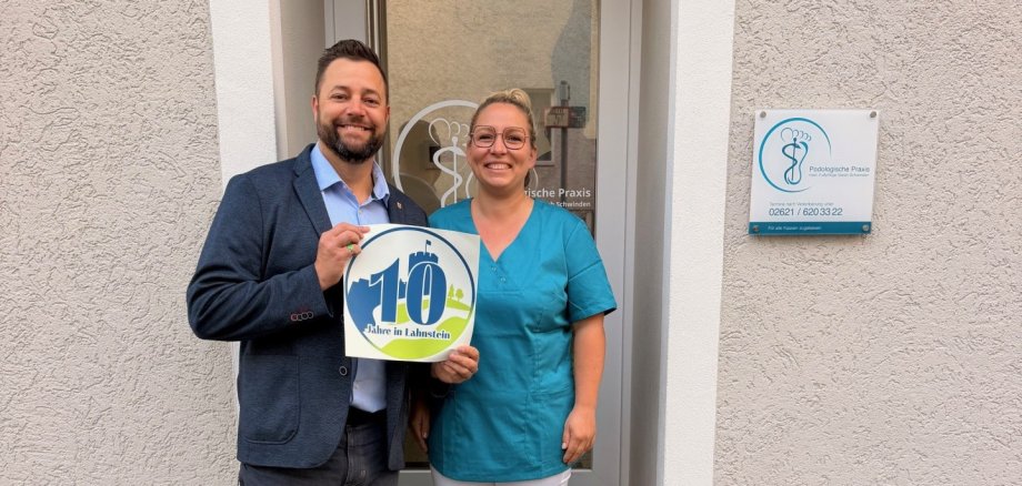 10 Jahre in Lahnstein: Oberbürgermeister Lennart Siefert überreichte Praxisinhaberin Sarah Schwinden das Lahnstein Label. Ein Mann und eine Frau stehen lächelnd vor dem Eingang eines Gebäudes und halten gemeinsam ein rundes Schild mit der Aufschrift „10 Jahre in Lahnstein“ in Blau, Grün und Weiß. Der Mann links trägt ein dunkelblaues Sakko, ein hellblaues Hemd, eine dunkle Hose und weiße Turnschuhe. Er hat kurze dunkle Haare und einen Bart. Die Frau rechts trägt eine türkisfarbene Tunika, eine weiße Hose und weiße Schuhe. Sie hat blonde, zusammengebundene Haare und trägt eine Brille. Hinter den beiden befindet sich eine gläserne Eingangstür mit weißem Rahmen. Rechts daneben hängt an der hell verputzten Hauswand ein Schild mit der Aufschrift „Podologische Praxis“ und einem stilisierten Fuß als Logo. Die beiden stehen auf und vor dunklen Steinstufen, die zu einem gepflasterten Gehweg führen.