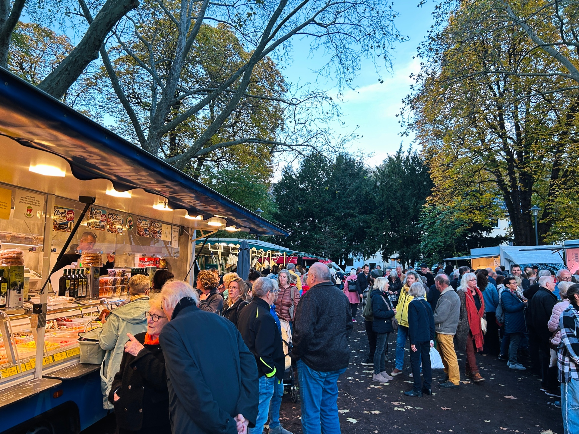 Zahlreiche Besucherinnen und Besucher auf dem gut besuchten Wochenmarkt im Schillerpark in Lahnstein am frühen Abend. Links im Bild steht ein großer, hell beleuchteter Verkaufswagen mit einer langen Glastheke, in der verschiedene mediterrane Spezialitäten wie Oliven, eingelegtes Gemüse und Feinkost angeboten werden. Mehrere Menschen stehen direkt vor der Theke und betrachten die Auslagen. Im Vordergrund unterhalten sich zwei ältere Personen, während sich dahinter zahlreiche weitere Marktbesucherinnen und -besucher zwischen den Verkaufsständen aufhalten. Rechts im Bild stehen Menschen in kleinen Gruppen zusammen oder schlendern über den Markt. Im Hintergrund sind weitere beleuchtete Marktstände zu sehen. Große Bäume mit teilweise gelb verfärbtem Herbstlaub umrahmen die Szene. Der Himmel ist noch hellblau, während die warmen Lichter der Verkaufsstände bereits für eine gemütliche Abendstimmung sorgen.