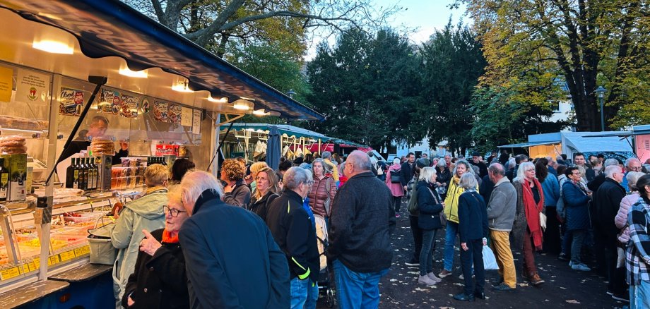 Zahlreiche Besucherinnen und Besucher auf dem gut besuchten Wochenmarkt im Schillerpark in Lahnstein am frühen Abend. Links im Bild steht ein großer, hell beleuchteter Verkaufswagen mit einer langen Glastheke, in der verschiedene mediterrane Spezialitäten wie Oliven, eingelegtes Gemüse und Feinkost angeboten werden. Mehrere Menschen stehen direkt vor der Theke und betrachten die Auslagen. Im Vordergrund unterhalten sich zwei ältere Personen, während sich dahinter zahlreiche weitere Marktbesucherinnen und -besucher zwischen den Verkaufsständen aufhalten. Rechts im Bild stehen Menschen in kleinen Gruppen zusammen oder schlendern über den Markt. Im Hintergrund sind weitere beleuchtete Marktstände zu sehen. Große Bäume mit teilweise gelb verfärbtem Herbstlaub umrahmen die Szene. Der Himmel ist noch hellblau, während die warmen Lichter der Verkaufsstände bereits für eine gemütliche Abendstimmung sorgen.