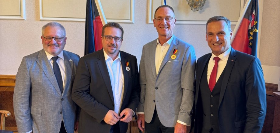 Vier Männer stehen nebeneinander in einem festlich eingerichteten Raum und lächeln in die Kamera. Der Mann ganz links trägt eine Brille, ein graues Sakko, ein weißes Hemd und eine dunkelblaue Krawatte. Neben ihm steht ein Mann mit Brille, dunklem Sakko, weißem Hemd, blauer Jeans und grauen Turnschuhen. An seinem Revers ist eine kleine Medaille mit schwarz-rot-goldenem Band befestigt. Der dritte Mann ist größer als die anderen, trägt eine Brille, ein hellgraues Sakko, ein weißes Hemd, eine dunkle Hose und braune Lederschuhe. Auch an seinem Sakko ist eine Medaille mit schwarz-rot-goldenem Band zu sehen. Rechts steht ein Mann in einem dunkelblauen dreiteiligen Anzug mit weißem Hemd und roter Krawatte. Im Hintergrund befinden sich zwei Fahnen in Schwarz-Rot-Gold und den Farben von Rheinland-Pfalz. Der Raum hat helle, mit Zierleisten verzierte Wände, eine Wandleuchte und einen hellen Parkettboden.
