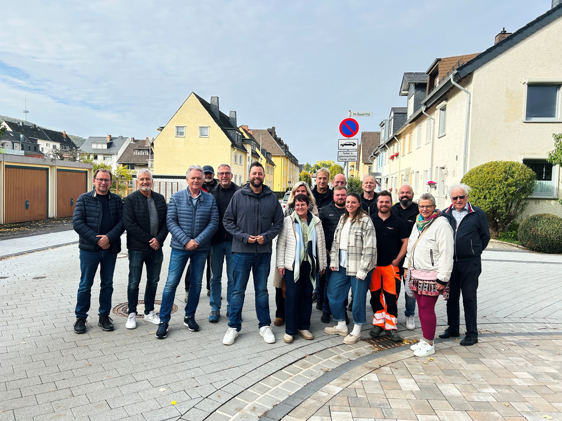 Eine Gruppe von 16 Frauen und Männern unterschiedlichen Alters steht für ein gemeinsames Gruppenfoto auf dem neu gepflasterten Lindenweg in Lahnstein. Die Personen stehen in zwei leicht versetzten Reihen nebeneinander und blicken überwiegend lächelnd in die Kamera. Sie tragen überwiegend Freizeitkleidung mit Jacken, Jeans und Turnschuhen. Ein Mann im vorderen rechten Bereich trägt eine schwarze Arbeitskleidung mit auffälliger orangefarbener Warnhose. Im Vordergrund ist die hellgraue und beigefarbene Pflasterung der Straße mit geschwungenen dunkleren Pflasterstreifen zu sehen. Im Hintergrund stehen Wohnhäuser mit hellen Fassaden, Garagen und vereinzelt geparkte Autos. Rechts hinter der Gruppe befindet sich ein Halteverbotsschild mit dem Straßenschild „Im Bauerntal“.
