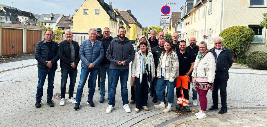Eine Gruppe von 16 Frauen und Männern unterschiedlichen Alters steht für ein gemeinsames Gruppenfoto auf dem neu gepflasterten Lindenweg in Lahnstein. Die Personen stehen in zwei leicht versetzten Reihen nebeneinander und blicken überwiegend lächelnd in die Kamera. Sie tragen überwiegend Freizeitkleidung mit Jacken, Jeans und Turnschuhen. Ein Mann im vorderen rechten Bereich trägt eine schwarze Arbeitskleidung mit auffälliger orangefarbener Warnhose. Im Vordergrund ist die hellgraue und beigefarbene Pflasterung der Straße mit geschwungenen dunkleren Pflasterstreifen zu sehen. Im Hintergrund stehen Wohnhäuser mit hellen Fassaden, Garagen und vereinzelt geparkte Autos. Rechts hinter der Gruppe befindet sich ein Halteverbotsschild mit dem Straßenschild „Im Bauerntal“.