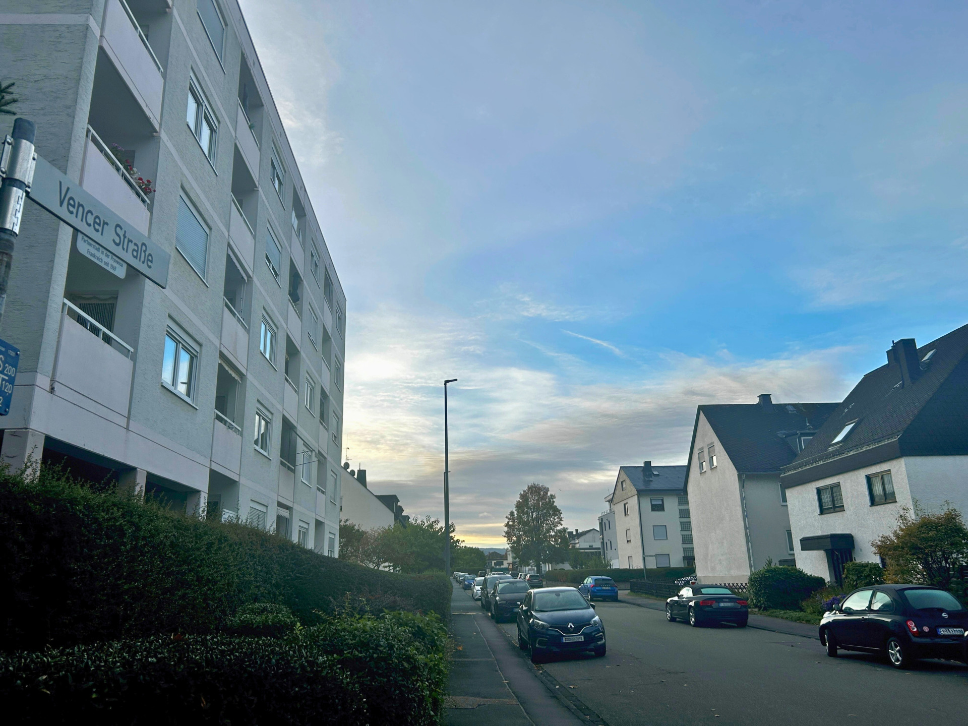 Blick entlang der Vencer Straße in Lahnstein mit parkenden Autos auf beiden Straßenseiten. Links steht ein mehrgeschossiges Wohngebäude mit Balkonen, rechts befinden sich mehrere Wohnhäuser. Im Vordergrund ist das Straßenschild „Vencer Straße“ zu sehen.