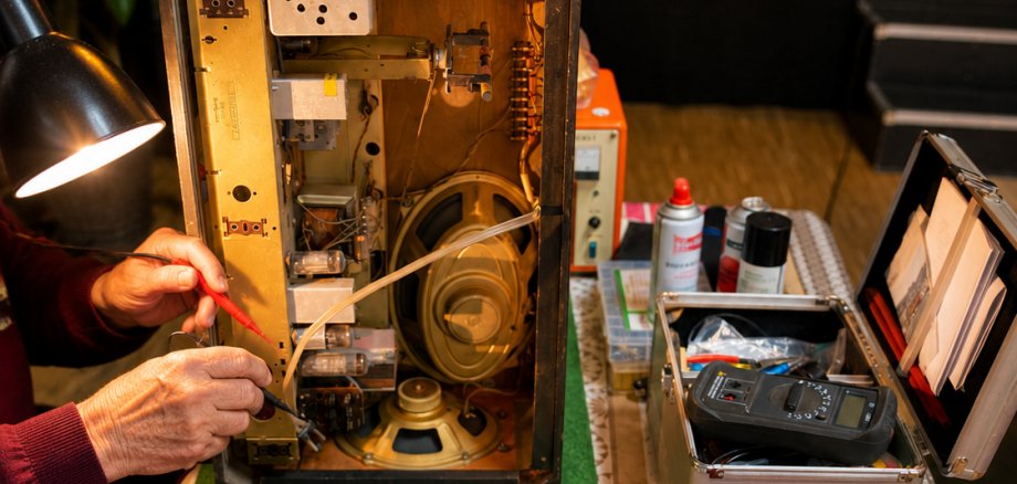 Eine Person überprüft mit Messgeräten das geöffnete Innenleben eines älteren Radios. Auf dem Arbeitstisch liegen verschiedene Werkzeuge und Geräte, darunter Multimeter, Schraubendreher und eine Lötstation.