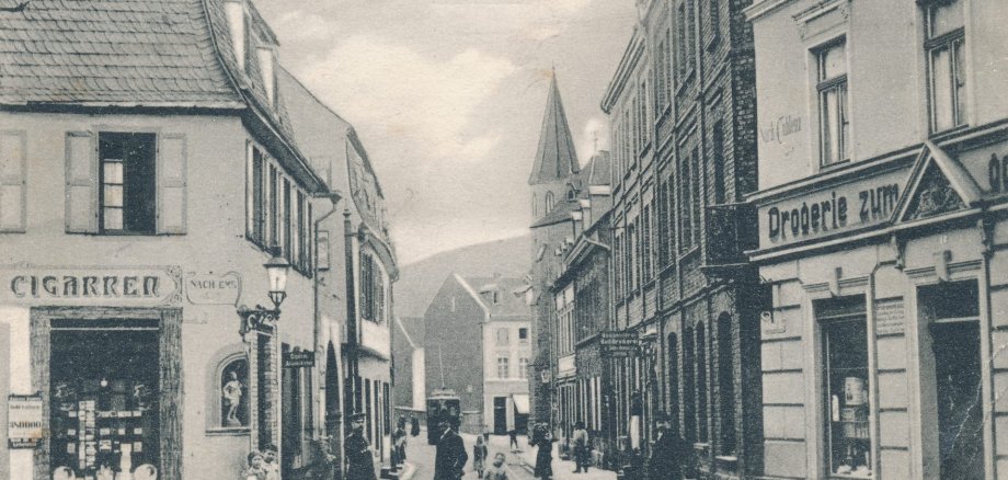 Erkennen Sie Lahnstein? Historische Aufnahme der Hochstraße aus dem Jahr 1907. Zu sehen sind zahlreiche Wohn- und Geschäftshäuser mit damaligen Ladenbeschriftungen. Mehrere Erwachsene und Kinder stehen auf der Straße; im Hintergrund erhebt sich ein Kirchturm.