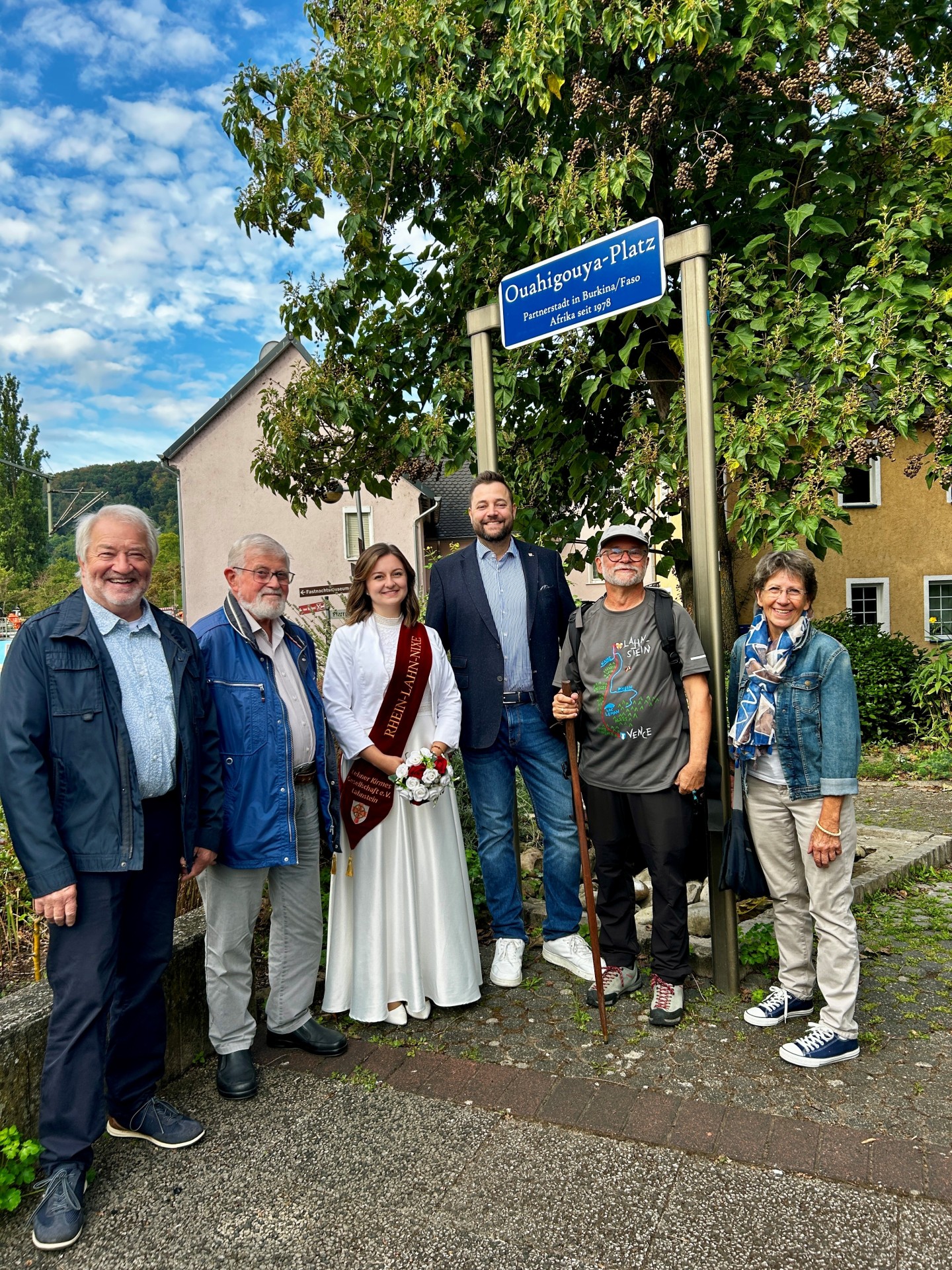 Sechs Personen stehen im Freien nebeneinander unter einem blauen Straßenschild mit der Aufschrift „Ouahigouya-Platz“. In der Mitte steht eine junge Frau in einem langen weißen Kleid mit einer rotbraunen Schärpe und einem kleinen Blumenstrauß. Neben ihr steht ein Mann in dunklem Sakko, Hemd, Jeans und weißen Schuhen. Rechts davon trägt ein Mann mit Brille und Kappe einen Rucksack und hält einen langen Wanderstock in der Hand. Die übrigen Personen tragen Freizeitkleidung. Im Hintergrund sind dichtes grünes Laub, Wohnhäuser und ein teilweise bewölkter blauer Himmel zu sehen.