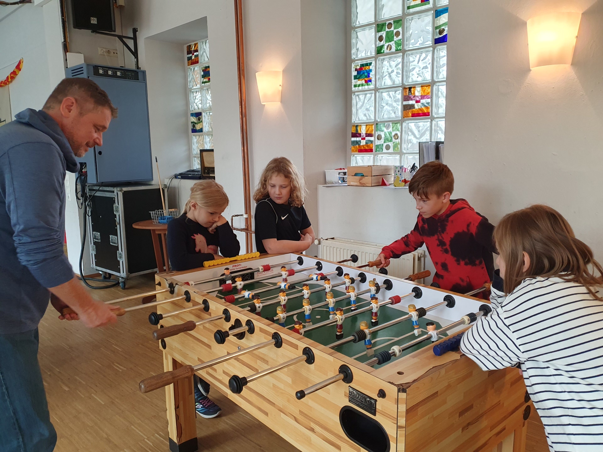 Ein Erwachsener und vier Kinder stehen gemeinsam an einem Tischkicker im Jugendzentrum. Zwei von ihnen spielen gegeneinander, während die anderen das Spiel aufmerksam verfolgen. Im hellen Raum sorgen farbig gestaltete Glasbausteine für bunte Akzente.