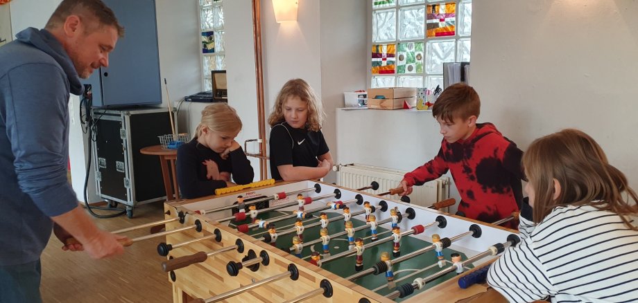 Kickerspielen im JUKZ Lahnstein Ein Erwachsener und vier Kinder stehen gemeinsam an einem Tischkicker im Jugendzentrum. Zwei von ihnen spielen gegeneinander, während die anderen das Spiel aufmerksam verfolgen. Im hellen Raum sorgen farbig gestaltete Glasbausteine für bunte Akzente.