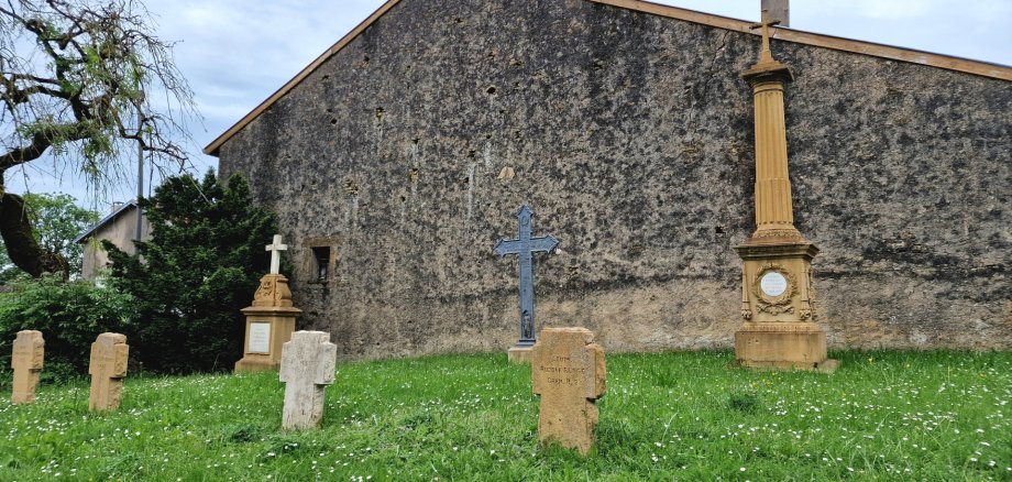 Kriegsgräber Kriegsgräber auf einer grünen Wiese vor einer hohen, alten Steinmauer. Mehrere schlichte Grabkreuze aus hellem und rötlichem Stein stehen verteilt auf der Fläche. Dazwischen befinden sich größere, aufwendig gestaltete Grabdenkmäler, darunter ein dunkles Metallkreuz und eine hohe steinerne Säule mit Kreuz. Die Wiese ist mit kleinen weißen Blüten bewachsen. Links ragt ein alter Baum ins Bild.