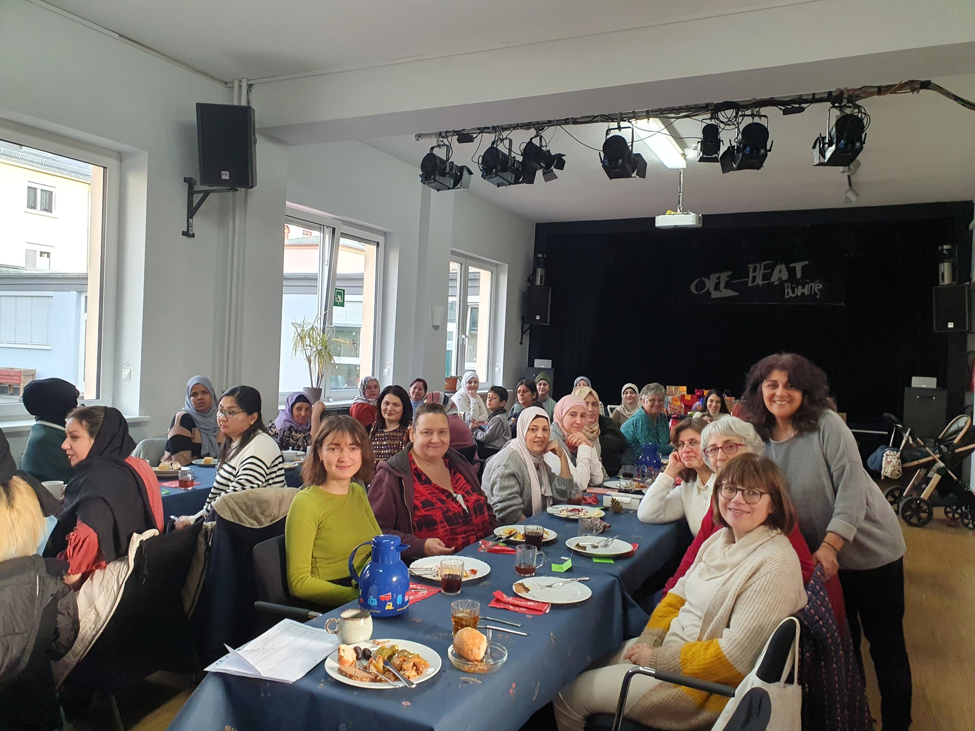 Eine größere Gruppe von Frauen sitzt gemeinsam an mehreren gedeckten Tischen in einem Veranstaltungsraum. Viele von ihnen blicken lächelnd in die Kamera. Auf den Tischen stehen Teller mit Speisen, Getränke und Kaffeekannen. Im Hintergrund befindet sich eine kleine Bühne mit Scheinwerfern.