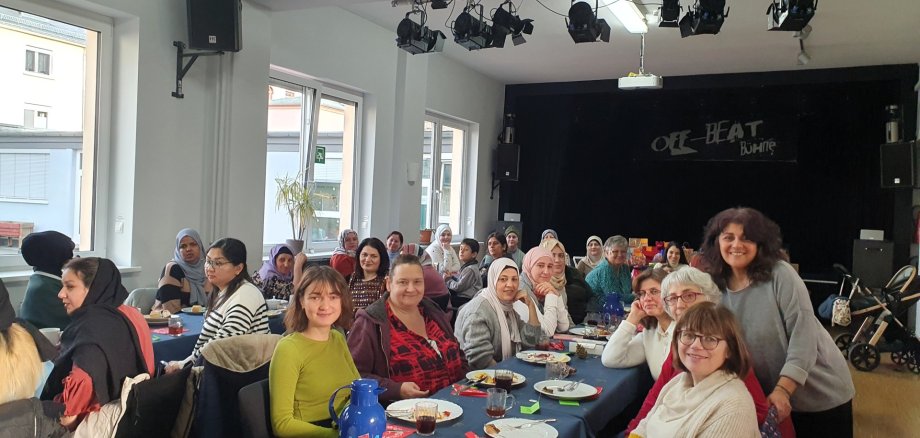 Interkulturelles Frauencafé im JUKZ Eine größere Gruppe von Frauen sitzt gemeinsam an mehreren gedeckten Tischen in einem Veranstaltungsraum. Viele von ihnen blicken lächelnd in die Kamera. Auf den Tischen stehen Teller mit Speisen, Getränke und Kaffeekannen. Im Hintergrund befindet sich eine kleine Bühne mit Scheinwerfern.