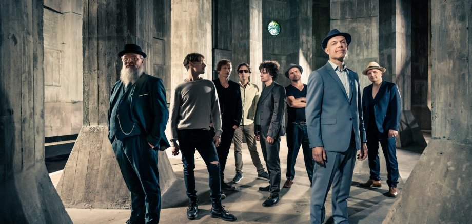 Thorbjørn Risager and The Black Tornado Acht Männer stehen in einer weitläufigen Halle zwischen hohen, massiven Betonpfeilern. Die Männer tragen überwiegend dunkle Kleidung, mehrere von ihnen Hüte. Zwei Männer stehen im Vordergrund, die übrigen sechs etwas versetzt dahinter. Durch seitlich einfallendes Licht entstehen deutliche Schatten auf dem Boden und den Betonflächen.