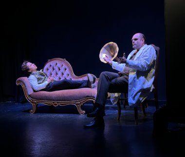 Zwei männliche Darsteller auf einer dunkel gehaltenen Theaterbühne. Links liegt ein Mann in Hemd und gemusterter Weste auf einer rosafarbenen, geschwungenen Chaiselongue. Rechts sitzt ein Mann mit weißem Kittel auf einem Holzstuhl und gestikuliert mit beiden Händen. Zwischen ihnen steht ein Grammophon mit großem Trichter. Die Szene ist in blau-violettes Bühnenlicht getaucht.