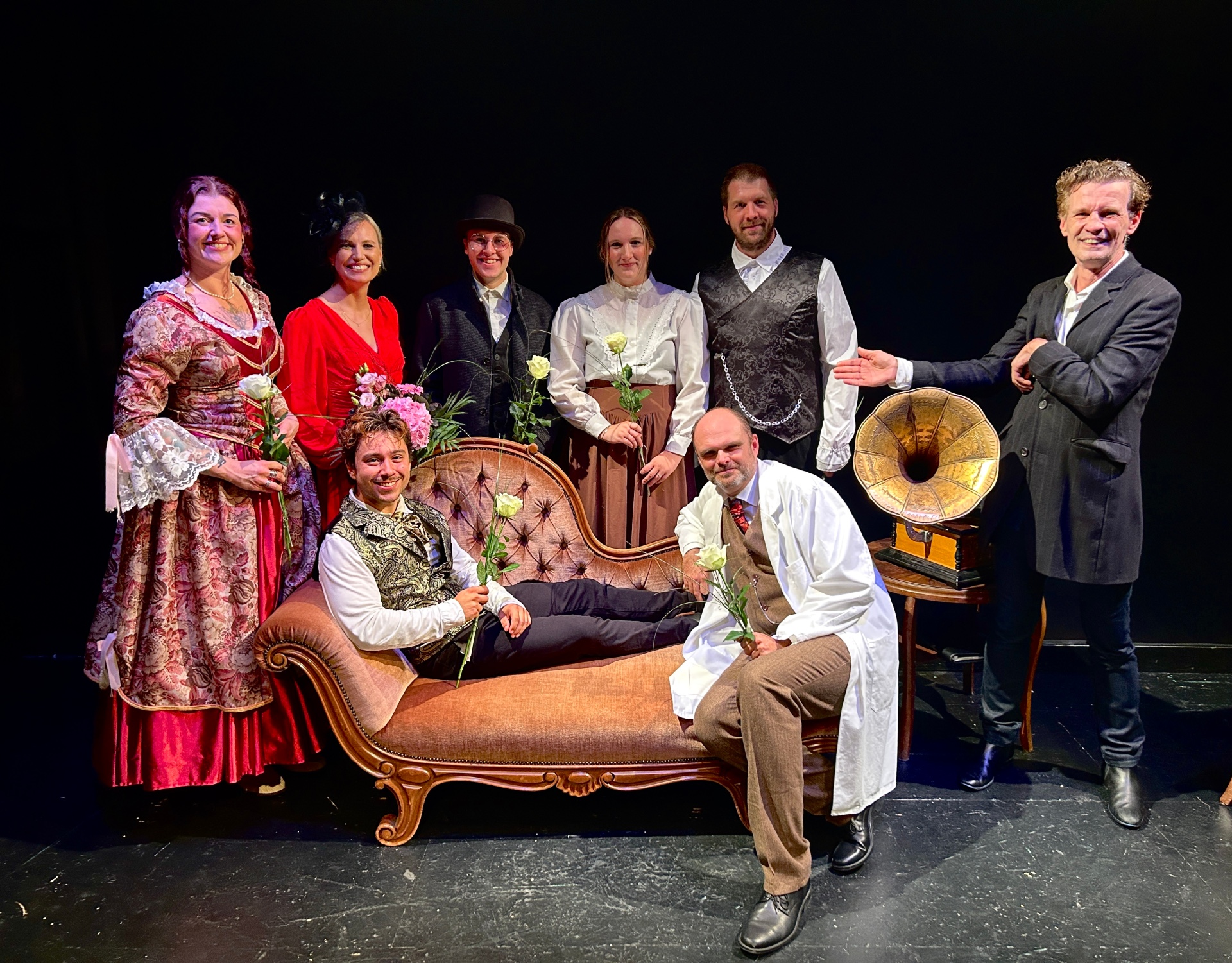 Gruppenaufnahme von neun Personen auf einer dunkel gehaltenen Theaterbühne. Acht von ihnen tragen historisch wirkende Kostüme, darunter lange Kleider, Westen, Hemden, einen weißen Kittel sowie verschiedene Hüte. Mehrere Personen halten weiße Rosen oder Blumensträuße. Ein Mann liegt vorne auf einer rosafarbenen Chaiselongue, ein weiterer kniet daneben. Rechts steht ein Mann in dunkler Jacke und deutet mit ausgestrecktem Arm auf die Gruppe. Neben ihm befindet sich ein Grammophon auf einem kleinen Holztisch.