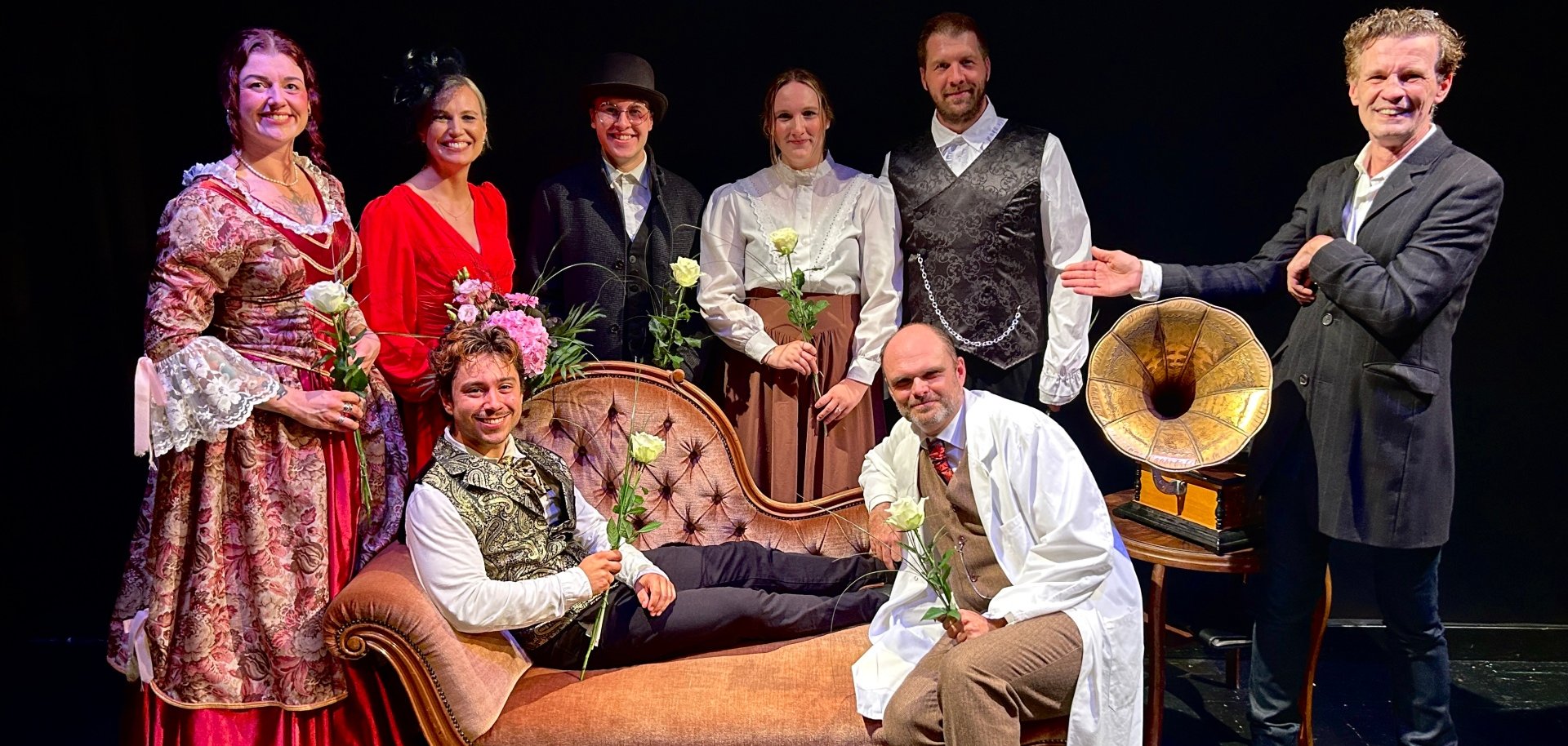 Gruppenaufnahme von neun Personen auf einer dunkel gehaltenen Theaterbühne. Acht von ihnen tragen historisch wirkende Kostüme, darunter lange Kleider, Westen, Hemden, einen weißen Kittel sowie verschiedene Hüte. Mehrere Personen halten weiße Rosen oder Blumensträuße. Ein Mann liegt vorne auf einer rosafarbenen Chaiselongue, ein weiterer kniet daneben. Rechts steht ein Mann in dunkler Jacke und deutet mit ausgestrecktem Arm auf die Gruppe. Neben ihm befindet sich ein Grammophon auf einem kleinen Holztisch.